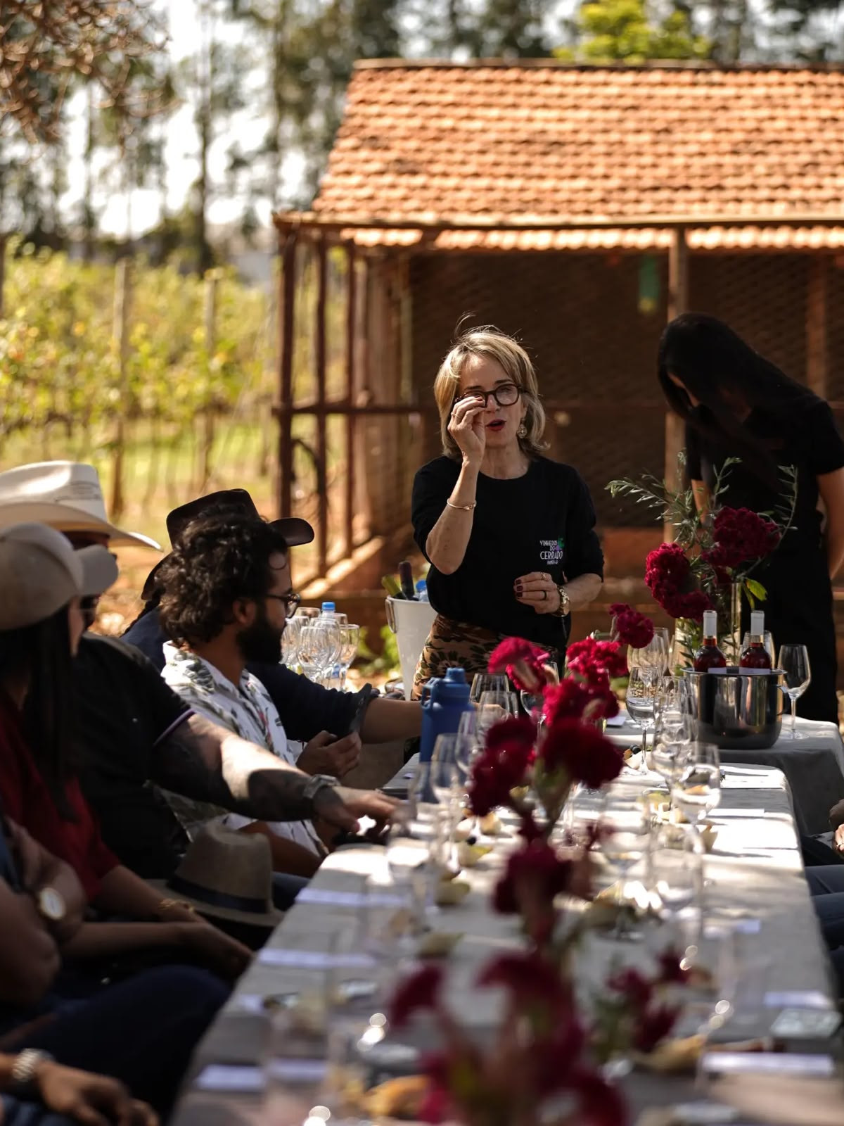 Anfitriã de camisa da vinícola apresenta os vinhos a visitantes sentados à mesa comprida, no parreiral