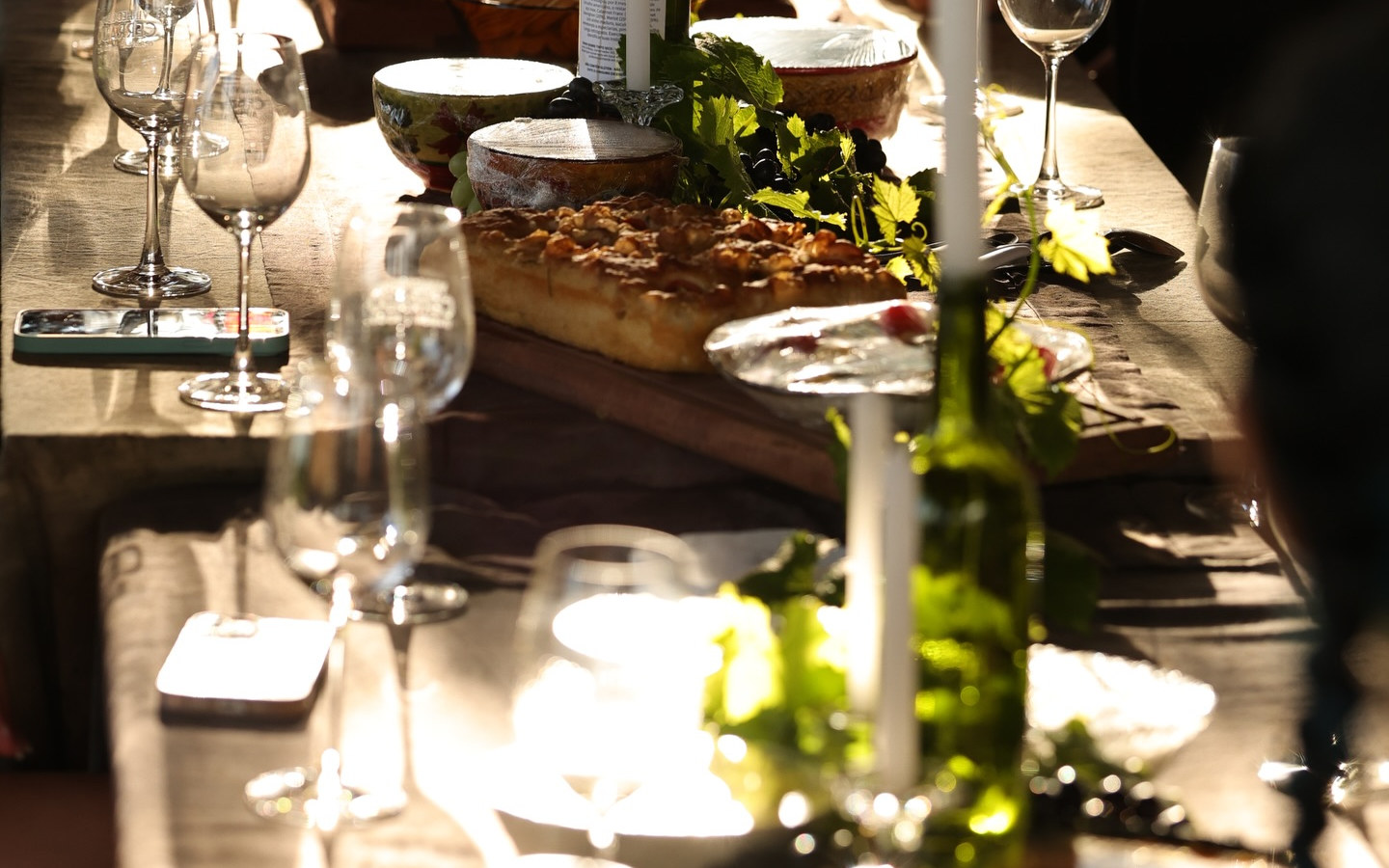 Mesa comunitária posta no parreiral ao entardecer, com pães, velas em garrafas de vinho e taças em contraluz