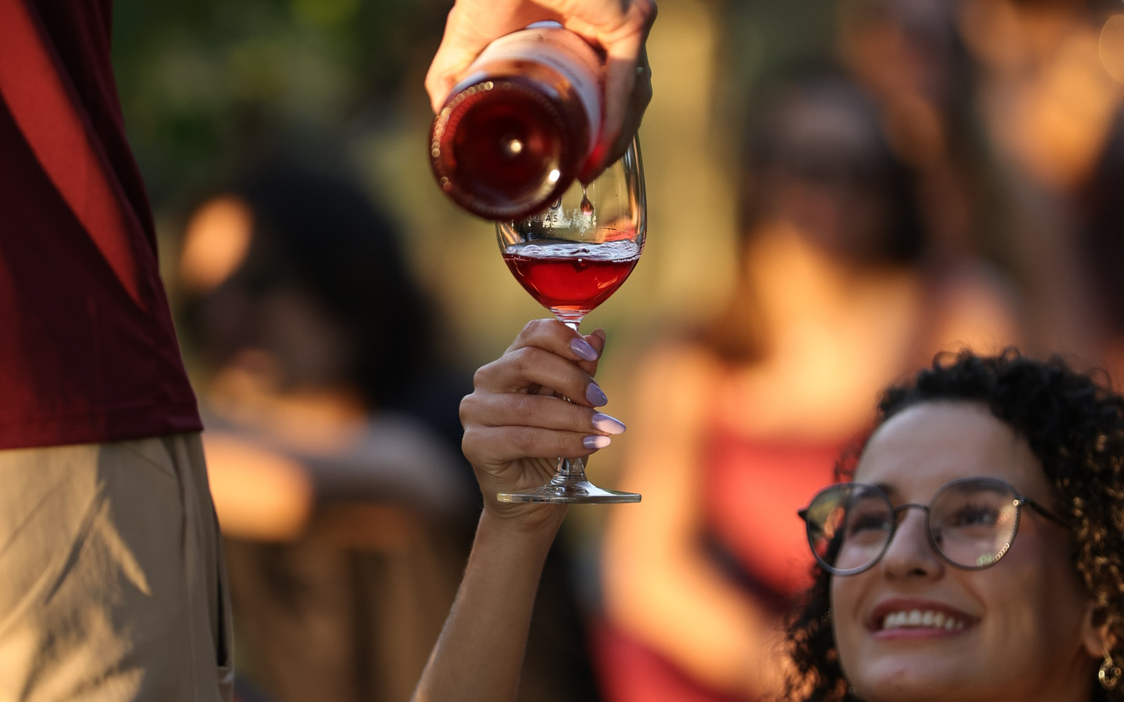 Convidada sorri e ergue a taça enquanto recebe vinho da garrafa, na luz dourada do fim de tarde